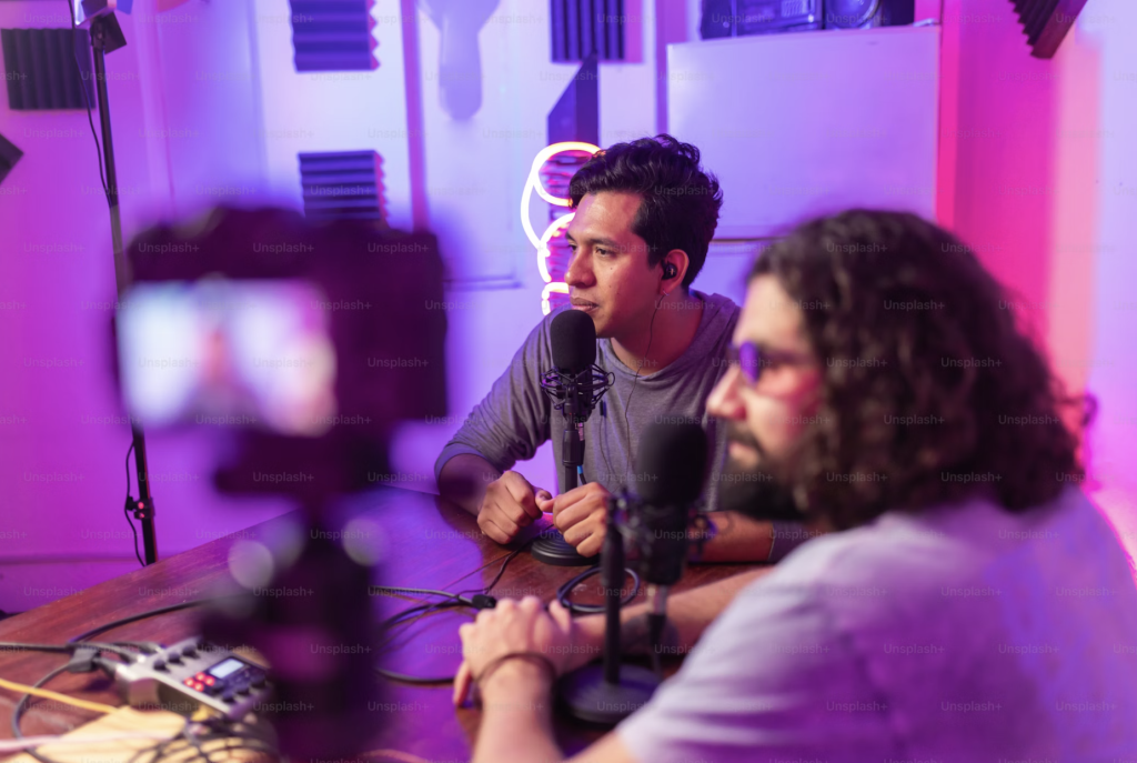 Two YouTube creators recording in a neon-lit studio, speaking into professional microphones with soundproof foam panels in the background.